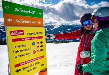 A winter sports enthusiast points at a signpost for the SkiWelt Tour in Hopfgarten. In the background, snow-covered mountains and a blue sky can be seen.