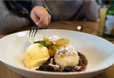 A plate with dessert, consisting of ice cream, a pastry, and a fruit sauce. In the background, hands and drinks are visible.