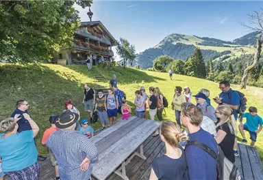 Eine Gruppe von Menschen versammelt sich im Freien vor einer Alm. Im Hintergrund sind grüne Hügel und ein blauer Himmel zu sehen.