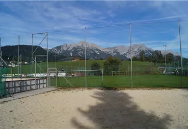 Ein Sandplatz vor einer Sportanlage mit Blick auf majestätische Berge. Der Himmel ist klar und die Umgebung ist grün und einladend.