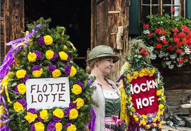 Eine Frau in traditioneller Kleidung hält zwei prächtig geschmückte Kränze. Im Hintergrund steht ein rustikales Holzgebäude mit blühenden Blumen.