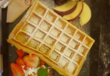 A delicious waffle with powdered sugar, served with fresh fruits like strawberries, apples, and grapes. Edible flowers and a serving of cream are also visible on the plate.