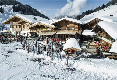 A cozy mountain cabin in the snow with many skis and winter sports enthusiasts. In the background, there are snowy mountains and a clear blue sky.