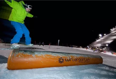 A snowboarder jumps over an orange rail at night. The slope is illuminated and provides a winter atmosphere.