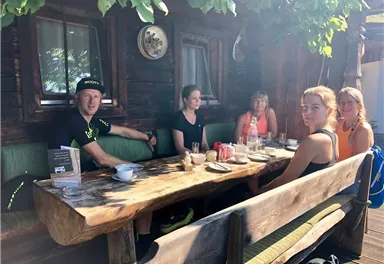 A group of five people is sitting at a large wooden table outdoors. They are enjoying drinks and snacks in a cozy, rural setting.