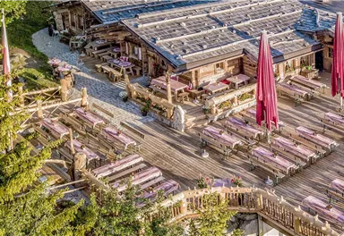 Ein gemütliches Restaurant mit einer großen Terrasse und vielen Sitzplätzen im Freien. Rote Sonnenschirme stehen zwischen den Tischen und umgeben von Bäumen.