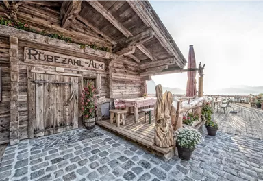 Eine rustikale Almhütte mit dem Namen "Rübezahl-Alm". Der Eingangsbereich ist mit Blumen und Holzverzierungen geschmückt.