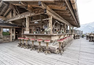 Eine gemütliche Berghütte mit einer Holzbar und mehreren Hockern. Im Hintergrund sind Berge zu sehen.