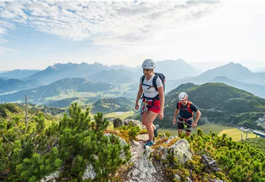 Two climbers are hiking on a mountain summit with a view of the surrounding mountains and the green valley. The landscape is surrounded by a clear, blue sky.