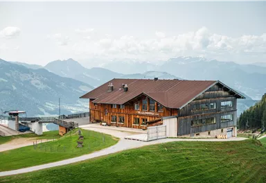 Ein großes Holzhaus auf einer Bergwiese mit Blick auf die umliegenden Berge. Der Himmel ist teilweise bewölkt und die Landschaft wirkt idyllisch und ruhig.
