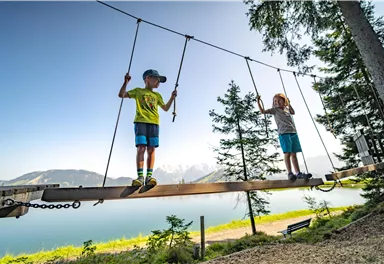 Two children are playing on a wobble bridge by the water. In the background, mountains and trees are visible.