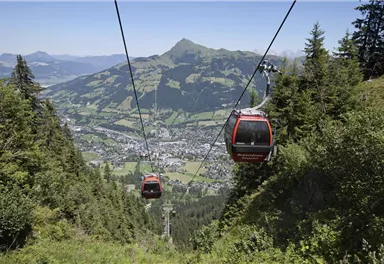 Eine Seilbahn fährt über eine grüne Landschaft mit Blick auf die Berge. Unten erstreckt sich ein malerisches Tal mit kleinen Häusern und viel Natur.
