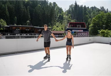 Ein Paar skatet Hand in Hand auf einer glatten Fläche. Im Hintergrund sind Bäume und Gebäude zu sehen.