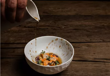 A handheld kettle pours broth over a colorful salad in a white bowl. The table is made of dark wood, creating a warm background.