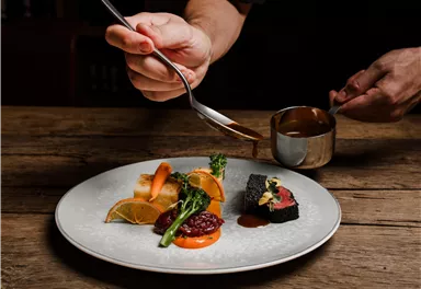 An artfully arranged plate with various dishes. A hand pours sauce over the dish on a wooden table.