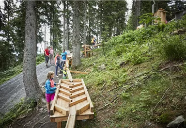 A group of people is hiking on a forest path with wooden structures. Surrounded by trees and dense vegetation, they enjoy nature.