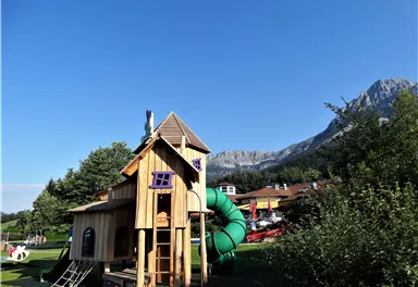 A playground with a wooden playhouse and a green slide. In the background, mountains and trees can be seen.