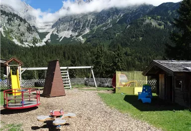 A playground surrounded by mountains and trees. The area features various play equipment and a sunny view.