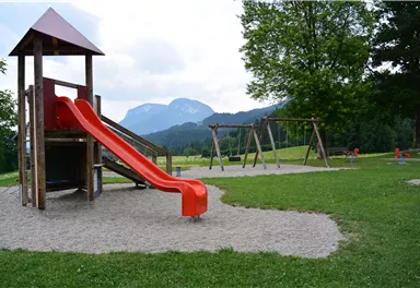 Ein Spielplatz mit einer roten Rutsche und Schaukeln. Im Hintergrund sind grüne Hügel und Berge zu sehen.