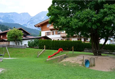 Ein Spielplatz mit einer Rutsche und einem Baum. Im Hintergrund sind schöne Häuser und Berge zu sehen.