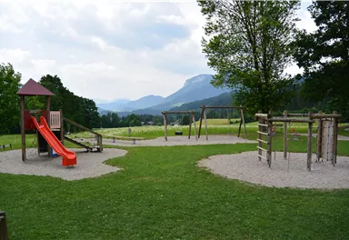 Ein Spielplatz in einer malerischen Umgebung mit einem Rutschturm und Schaukeln. Im Hintergrund sind Berge und eine grüne Wiese zu sehen.