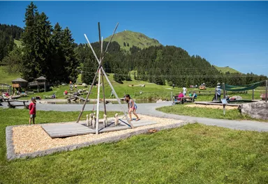 Ein Spielplatz in einer schönen, grünen Landschaft mit Hügeln im Hintergrund. Kinder genießen die verschiedenen Spielmöglichkeiten unter einem klaren blauen Himmel.