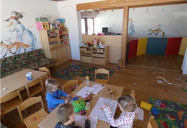 Ein kunterbunter Spielraum für Kinder mit Tischen, Stühlen und vielfältigen Bastelmaterialien. An den Wänden sind fröhliche Zeichnungen und ein Spielbereich mit bunten Trennwänden.