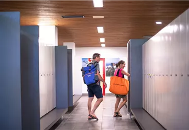 Zwei Personen gehen in einem modernen Umkleideraum vorbei. Die Umgebung hat Schließfächer und eine helle Beleuchtung.
