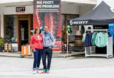 Two people are standing in front of a store with outdoor gear. In the background, there are advertising banners and sales booths visible.