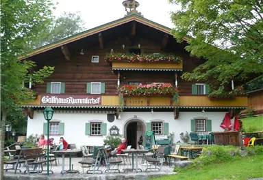 Ein traditionelles Holzhaus mit blühenden Balkonen und einer einladenden Terrasse. Gäste sitzen draußen und genießen die ruhige Umgebung.