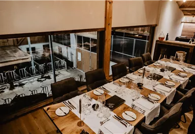 Ein elegantes Restaurant mit einem großen Tisch, der für ein Essen gedeckt ist. Große Fenster bieten einen Blick auf die Outdoor-Bereiche.