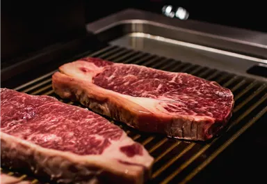 Saftige Steaks liegen auf dem Grill. Im Hintergrund sind Hände zu sehen, die sich auf das Grillen vorbereiten.