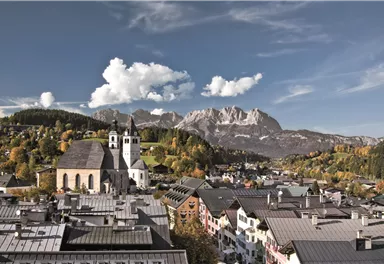 A picturesque town with a historic church tower and mountains in the background. The landscape is surrounded by colorful trees and the sky is partly cloudy.
