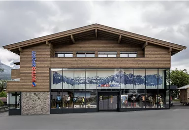 Ein modernes Gebäude mit Holzverkleidung und großen Fenstern. Im Hintergrund ist ein Bergmotiv zu sehen.