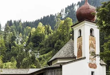 A charming church with a red onion dome stands among green trees. In the background, mountains and a cable car are visible.