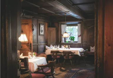 A cozy dining room with wooden walls and warm lighting.
Tables are set, and a window overlooking plants creates an inviting atmosphere.