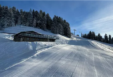 Ein winterliches Landschaftsbild mit einem Haus auf einem schneebedeckten Hang. Im Hintergrund sind Bäume und ein Skilift sichtbar.