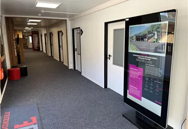 A bright hallway with a modern information screen and several doors. The floor is carpeted and there is a red seating area.