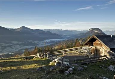 A picturesque mountain landscape with gentle hills and a view of a valley. In the foreground, there is a rustic cabin.