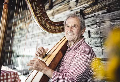 An older man is playing the harp and smiling. In the background, a rustic wooden wall is visible.