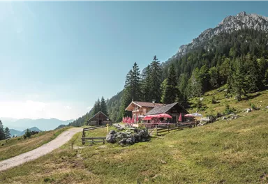 A picturesque mountain hut surrounded by green meadows and tall trees. In the background, impressive mountains rise under a clear sky.