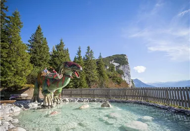 A large dinosaur stands in a crystal-clear water basin, surrounded by trees and mountains. The sky is blue and sunny.