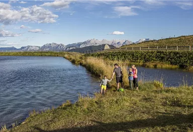 A family stands on the shore of a calm body of water, surrounded by nature and gentle hills. The mountains in the background are visible under a clear sky.