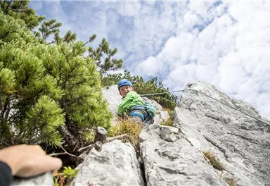A child is climbing on a rock wall and wearing a helmet. The surroundings are characterized by green plants and a cloudy sky.