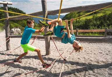 Two children are playing on a playground. One is hanging from a rope while the other is holding it.