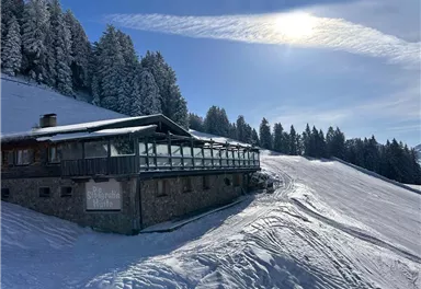 Eine idyllische Berghütte im Schnee, umgeben von verschneiten Bäumen. Der Himmel ist klar und die Sonne scheint.