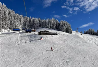 Eine schneebedeckte Landschaft mit einer Skihütte und Lifts. Klare blaue Himmel und verschneite Bäume im Hintergrund.