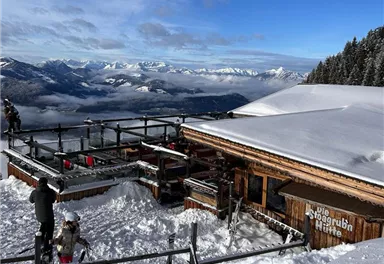 Eine Berghütte umgeben von Schnee mit Blick auf die Berge. Die Atmosphäre ist klar und sonnig.