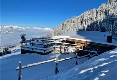 Ein verschneites Berghaus umgeben von schneebedeckten Bäumen. Der Himmel ist klar und blau, mit Blick auf die Berge in der Ferne.