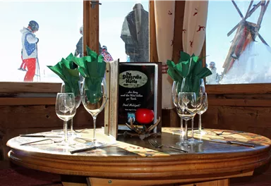 Ein gemütlicher Tisch in einer Berghütte mit Gläsern und Dekoration. Im Hintergrund sind Skifahrer zu sehen.